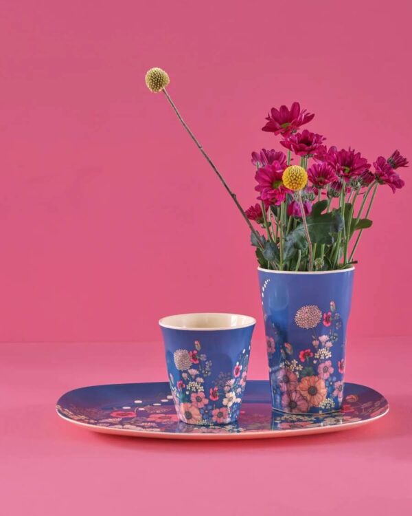 Rice Melamin Becher Größe L mit blauem Flower Collage Print fotografiert vor pinkem Hintergrund mit kleinem Becher und einem länglichen Teller des gleichen Designs und einem Strauß pinker Blumen