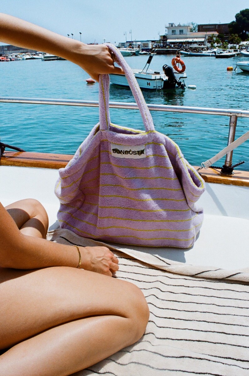 Bongusta Weekendbag in Lila mit gelben Streifen und Markenpatch auf der Vorderseite am Meer